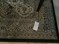 Photo of area rug showing black, beige, and brown floral pattern with a decorative border and a tag on the rug.