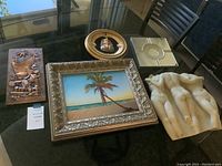 Photo of miscellaneous art pieces on a glass table including a framed beach scene painting, bronze relief plaque, circular decorative plate, white sculptural wall art, and beige tile ashtray.