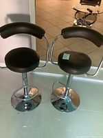 Photo shows two black and chrome bar stools with round cushioned seats and curved backrests, chrome pedestal bases, and footrests. Bar stools are placed on tiled floor.
