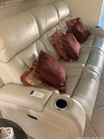Light beige or cream colored leather or faux leather three seat powered reclining sofa with maroon pillows and cup holder.