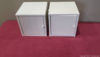Two white square metal wall cabinets with closed doors on red surface