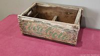 Full view of the antique wooden Coca-Cola crate showing the rectangular shape, open top, wooden divider inside, and faded green branding on side.