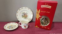 The infant's set including a small plate, bowl, and cup displayed next to original red Bunnykins box.
