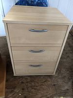 Front view of the three drawer wooden cabinet on caster wheels, showing the metal handles and wood grain.