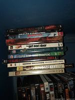 Stack of assorted DVD movies visible from top down, several titles readable including The Last House on the Left unrated, Juice, The Girl Next Door, and sports related titles.