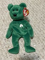 Green plush bear with black eyes and nose, white shamrock embroidery on chest, Ty heart tag on ear, lying on carpet.