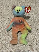 Front view of a tie-dye multi-colored TY Beanie Baby Peace Bear with red heart tag showing TY logo and embroidered peace sign on chest.