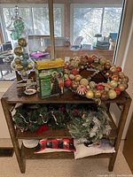 Wide view of wooden table filled with Christmas wreath decorated with ornaments, decorative pillows, a green box for tree dazzler pillows, glass vase with ornaments, and more decor items