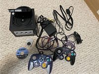 Black Nintendo GameCube console with power adapter, two wired controllers (one purple and one black), memory card, and LEGO Star Wars II game disc in the setup on carpet surface.