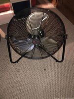 Front view of large black metal fan showing three silver blades, protective wire grill, and metal frame stand.