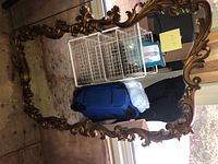 Large ornate mirror laying flat showing the full gold decorative frame with detailed scroll and floral patterns.