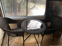 Patio conversation table and two chairs placed indoors near windows, visible outdoor view. Table is round with glass top, chairs are woven wicker style.