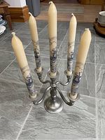 Full view of five-arm pewter candelabra holding five decorative taper candles placed on tile floor.