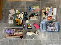 Wide view of sewing supplies and tools arranged in containers and trays on floor showing spools of thread, buttonholer, electric scissors, and assorted notions.