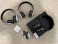 Photo shows two wireless over-ear headphones, one wired over-ear headphone with volume control, a pair of white wired earbuds in case, and two black cables, arranged on carpeted floor with one headphone on a black box.