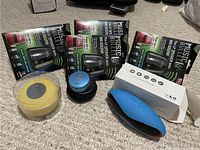 Four black Music Bullet wireless mini speakers in green and black box packaging along with one blue wireless speaker with red trim on carpeted floor.