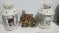 Front view of two white star-pattern metal lanterns and a miniature rustic house model placed between them.