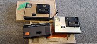 Photo showing three vintage cameras (Kodak Disc 6000 and two Winner pocket cameras) laid on floor with original box under one camera