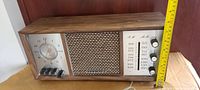 Front view of vintage RCA Victor radio showing wood casing, analog clock, speaker grille, AM/FM band display, and control knobs with measuring tape for size reference.
