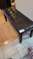 Front angle of dark brown leatherette cushioned bench with button tufting and dark wood legs on tile and wood floor.