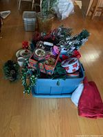 Two Rubbermaid containers filled with assorted Christmas decorations including faux plants, ribbons, cookie tins, ornaments, and holiday-themed items.