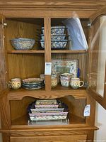 Photo of ceramic bowls, small square casseroles with lids, and cups arranged inside a wooden cabinet.