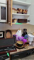Open cabinet showing multiple brown-toned ceramic mugs, plates and bowls, stacked, along with heat-resistant glass bowls with purple lids, knife set in wooden block, wooden cutting boards, water jug with filter, spice rack, and pepper grinder on counter.
