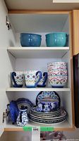 Full cabinet shelf with assorted dishware including turquoise mixing bowls on top shelf, floral mugs and bowls mid shelf, and stacked plates and teapot on bottom shelf.