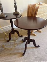 Pair of dark wood round pedestal end tables shown side by side on beige carpet, with folded wooden trays in background.