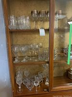 Glass cabinet showing multiple shelves of clear crystal glassware including stemmed glasses with etched detail, decorated wine glasses with gold and purple rims, and a large crystal vase on right side of bottom shelf.
