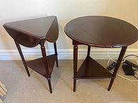 Pair of Bombay Company occasional tables. Includes one round table with three drop leaves and a triangular lower shelf, alongside a smaller triangular table with drop leaves on two sides and lower shelf. Both tables are dark wood.