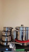 Stack of multiple stainless steel pots and pans on a black shelf with whisk in plastic bag