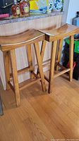 Two wooden kitchen stools positioned next to a counter, showing curved seats and four legs with horizontal supports, on wood floor.