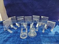 Ten clear Lucite display stands arranged on dark blue fabric. Various shapes including plate stands and cup holders, some with visible wear and minor surface scratches.