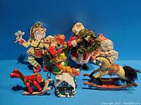 Seven assorted vintage Christmas decorations on blue background including Santa Claus figurines, a wooden rocking horse, a trinket box, and various ornaments.