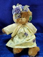 Front view of the 15 inch Bearington Bear teddy bear dressed in a floral dress and a hat with purple flowers on a blue background.