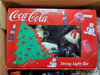 Boxed Coca Cola string light set with visible polar bear and Dalmatian figurines inside the packaging, showing branding and festive graphics.