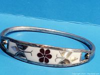 Close-up image showing inlaid floral and geometric design with abalone and white material on silver bangle bracelet.