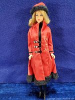 Full body image of the Barbie doll wearing the red velvet coat trimmed with black faux fur and black knee-high boots against a blue background.