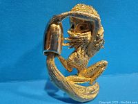 Front view of a vintage gold-tone brooch illustrating a person sitting and pouring water from a vessel, showing detailed textured gold metal and green eyes.