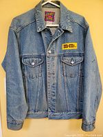 Front view of the blue denim jacket hanging on a hanger, showing button closure, two button flap chest pockets, and embroidered M&M's patch on left chest.