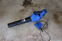 Blue Kobalt 24V Max brushless cordless leaf blower lying on concrete floor alongside blue battery charger with attached power cord. Battery is installed in the blower body.