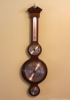 Full view of the Howard Miller 612-718 Catalina banjo wall clock and weather station hanging on the wall, showing the entire 31 inch tall wood frame with thermometer, clock, barometer, and hygrometer.