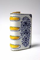 Front side view of the ceramic book flask showing hand-painted blue floral design, yellow decorative accents, and cork stopper on top.