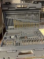 Large gray plastic case open displaying multiple rows of drill bits of various sizes and types. Some missing bits visible in empty slots.