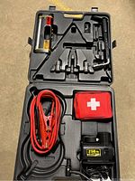 Opened black plastic case showing interior with jumper cables, first aid kit, air compressor and lug wrench arranged in molded compartments.