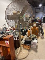 Photo of large industrial tall shop fan with metal cage and blades, mounted on tall stand with round base, in a warehouse or workshop environment.