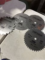 Photo shows four circular saw blades placed on a white surface. Each blade is marked with Craftsman branding and diameter of 7 1/4 inches. Teeth counts visible on some blades are 40T and 20T.