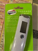 Front view of the American Tourister luggage scale in packaging showing the scale device, display screen, and button, with text about maximum weight and battery.
