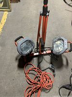 Orange metal tripod supporting two gray caged work lights, black handles, with orange extension cord coiled on the ground below